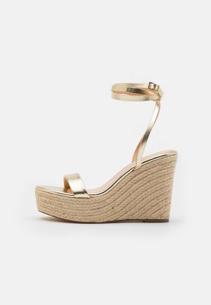 Sandale cu toc wedge din metalizat auriu, cu o bază din jutu țesut, având o curea lată pentru vârf și o curea pentru gleznă, textură netedă și un design elegant.