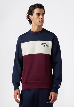 Joven luciendo un sudadera de Champion con bloques de color en navy, blanco y burdeos, mirando hacia su izquierda frente a un fondo blanco liso.
