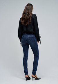 Kobieta o długich brązowych włosach, ubrana w czarną bluzkę, ciemnoniebieskie obcisłe dżinsy i czarne szpilki, stojąca na tle jednolitego szarego tła.