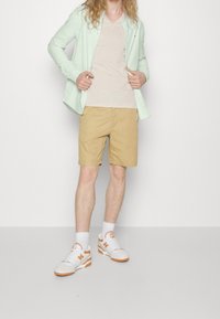 Ljus mintgrön skjorta med knapp-up över en beige V-ringad t-shirt, khakishorts och vita sneakers med orangea accenter. Släta, avslappnade tygstrukturer.