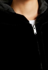 Chaqueta de pelo sintético negra con capucha. Presenta un cierre de cremallera plateado, un forro interior suave y una textura suave y esponjosa. Primer plano de la zona del cuello.