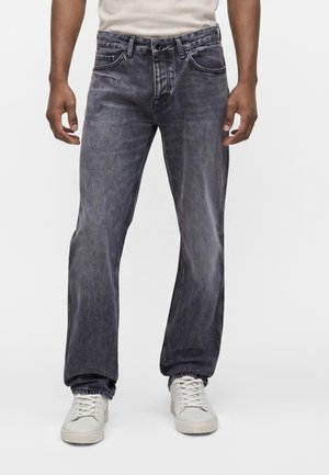 Man draagt vervaagde zwarte straight-leg jeans en witte sneakers, staand tegen een effen lichte achtergrond.
