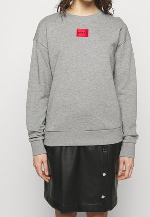 Person trägt einen grauen Pullover mit rotem "HUGO"-Aufnäher und einen schwarzen, mit Knöpfen versehenen Lederrrock, steht vor weißem Hintergrund.