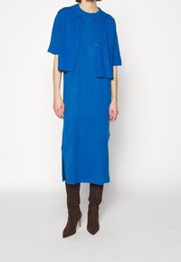 Blå stickad klänning med krage och kortärmad överdel, med sidofickor och bärs tillsammans med bruna knähöga stövlar. Mjuk textur, enkel design.