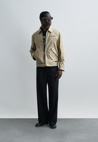 Άνδρας που φοράει μπεζ ζακέτα με φερμουάρ, μαύρο παντελόνι με φαρδύ πόδι, μαύρα παπούτσια και σκούρα γυαλιά ηλίου, στέκεται μπροστά σε απλό γκρι φόντο.