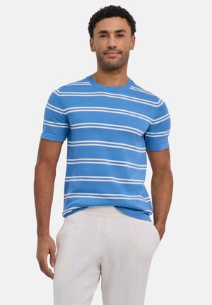 Man met een blauwe, korte mouw gebreide trui met witte horizontale strepen en lichtgekleurde broek, staand met één hand in de zak.