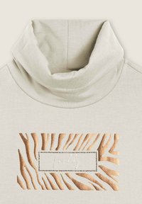 Top de cuello tortuga beige claro, que presenta un estampado de cebra texturizado en dorado y marrón, con un logo central en blanco escrito a mano. Tejido suave y casual.