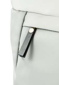 Angolo di uno zaino in tessuto grigio con cerniera nera, dotato di un pulsante accentato rotondo in argento e dettaglio testurizzato sul tirante della cerniera.