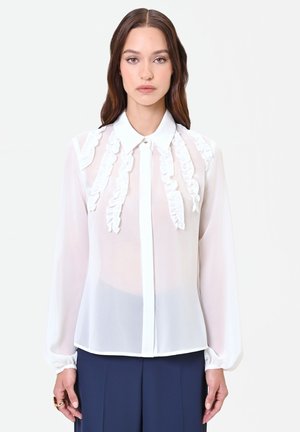 Frau trägt eine transparente weiße Bluse mit Rüschen-Details vorne, kombiniert mit dunkelblauen Hosen, steht vor einem einfarbigen Hintergrund.