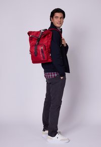 Czerwona plecak z regulowanymi paskami, przednimi zamkami i miękką fakturą; noszony przez osobę w granatowej kurtce i ciemnych spodniach.
