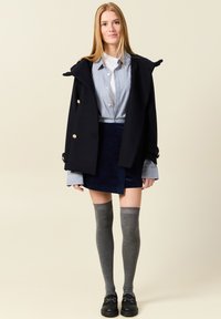 Cappotto doppiopetto navy con bottoni dorati, indossato sopra una camicia a righe blu e bianche, abbinato a una gonna mini navy e calze al ginocchio grigie.