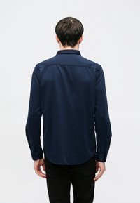 Navyblauw shirt met lange mouwen gemaakt van gladde stof; heeft een klassieke kraag en knoopmanchetten, met een afgeronde zoom aan de achterkant.