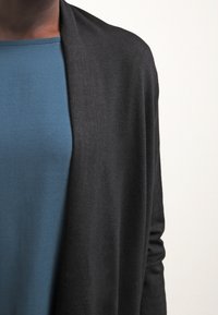 Černý kardigan s otevřeným předním dílem, vyrobený z měkké, lehké látky. Nositelný přes hladké, přiléhavé tyrkysové tričko. Jednoduchý, minimalistický design.