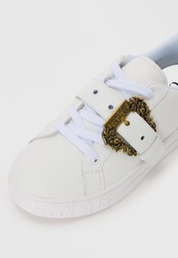 White sneaker with a smooth leather upper, featuring a gold decorative buckle and white laces. Textured sole has an embossed brand name.