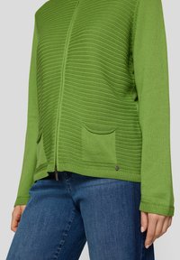 Grüne Strickjacke mit strukturiertem Muster, Reißverschluss und zwei Vorderentaschen. Kombiniert mit blauen Denim-Jeans.