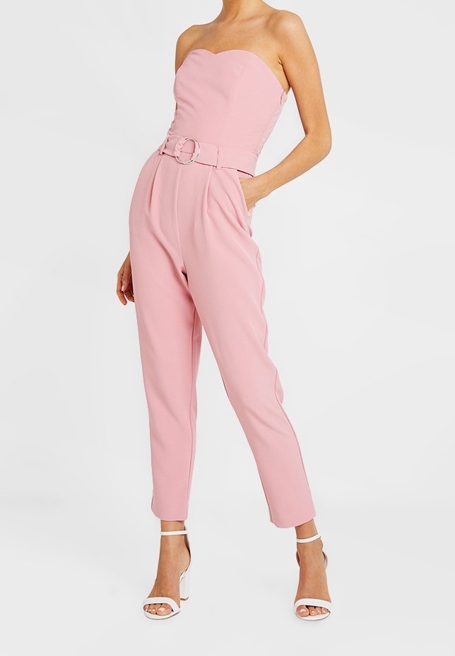 Combinaison rose sans bretelles avec un corsage ajusté et des jambes larges. Dispose d'une ceinture avec une boucle circulaire et d'une texture lisse. Fond uni.