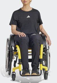 Juodas sportinis marškinėliai su balta logo, juodos kelnes ir juodais sportbačiais. Asmuo sėdi geltonoje invalidų vežimėlyje su dideliais ratais.