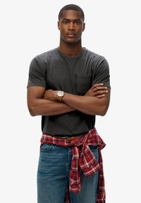 Grijze t-shirt met korte mouwen en zak, blauwe jeans en een vastgebonden rood geruit overhemd om de taille, met een gouden horloge om de pols.