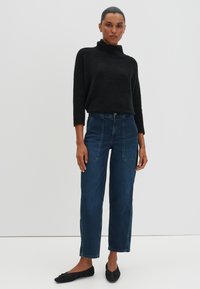 Maglione a collo alto nero con texture, jeans blu scuro a vita alta con tasche a toppa e ballerine nere, design minimalista su uno sfondo neutro.