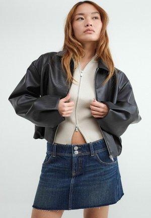 Jeune femme portant une veste en cuir noire sur un pull beige à fermeture éclair devant et une mini-jupe en jean bleue, posant devant un fond uni.