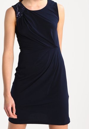 Robe bleu foncé sans manches avec un drapé latéral et un détail pailleté sur l'épaule gauche, portée par une personne au bras détendu.