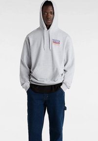 Grijze hoodie met verstelbare trekkoord, voorzak en blauw "VANS" logo op de borst. Draagt over een zwart shirt. Gecombineerd met denim broek.