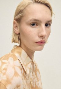 Modella bionda con capelli corti che indossa una camicia button-up beige con un motivo. Il tessuto presenta leggeri disegni floreali e una trama morbida.