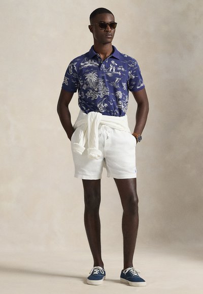 Άνδρας που φοράει μπλε σκούρο πουά πόλο, λευκές κοντές βερμούδες, λευκό πουλόβερ δεμένο στη μέση, μπλε σκούρα αθλητικά παπούτσια, γυαλιά ηλίου και ρολόι χειρός, στέκεται μπροστά σε απλό φόντο.