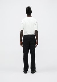 Man die met de rug naar een effen muur staat, gekleed in een wit shirt met korte mouwen, zwarte broek en zwarte schoenen, met ontspannen handen langs het lichaam.