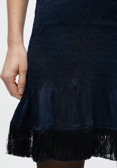 Tengerészkék ruha texturált smockkal, fodros szegéllyel, amely fekete bojtokkal díszített. Kéz fogja meg a szegélyt a részletekért.