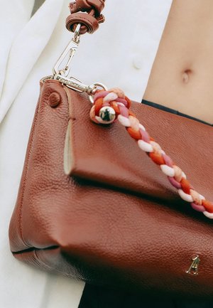 Sac à main en cuir marron avec un détail de sangle tressée rose, orange et blanche, porté par une personne vêtue de blanc qui montre le ventre.