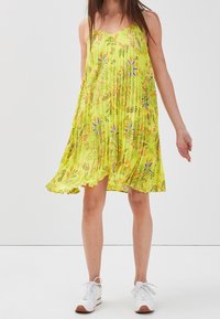 Robe sans manches jaune avec un design plissé, motifs floraux en bleu et orange, et une jupe évasée. Portée avec des baskets blanches.