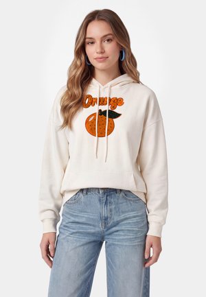 Ung kvinde med langt bølget hår iført blå hoop-øreringe, en hvid hættetrøje med orange grafik og tekst samt lyseblå jeans.