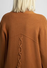 Sweter w kolorze rdzy o miękkiej fakturze. Charakteryzuje się charakterystycznym tylnym wzorem z krzyżującymi się sznurkami materiału wzdłuż kręgosłupa.