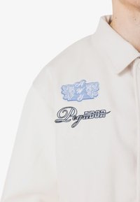 Hvit skjorte med krage-design. Inkluderer blå broderte detaljer, inkludert et mønstrete logo og teksten "Редактор" i svart.