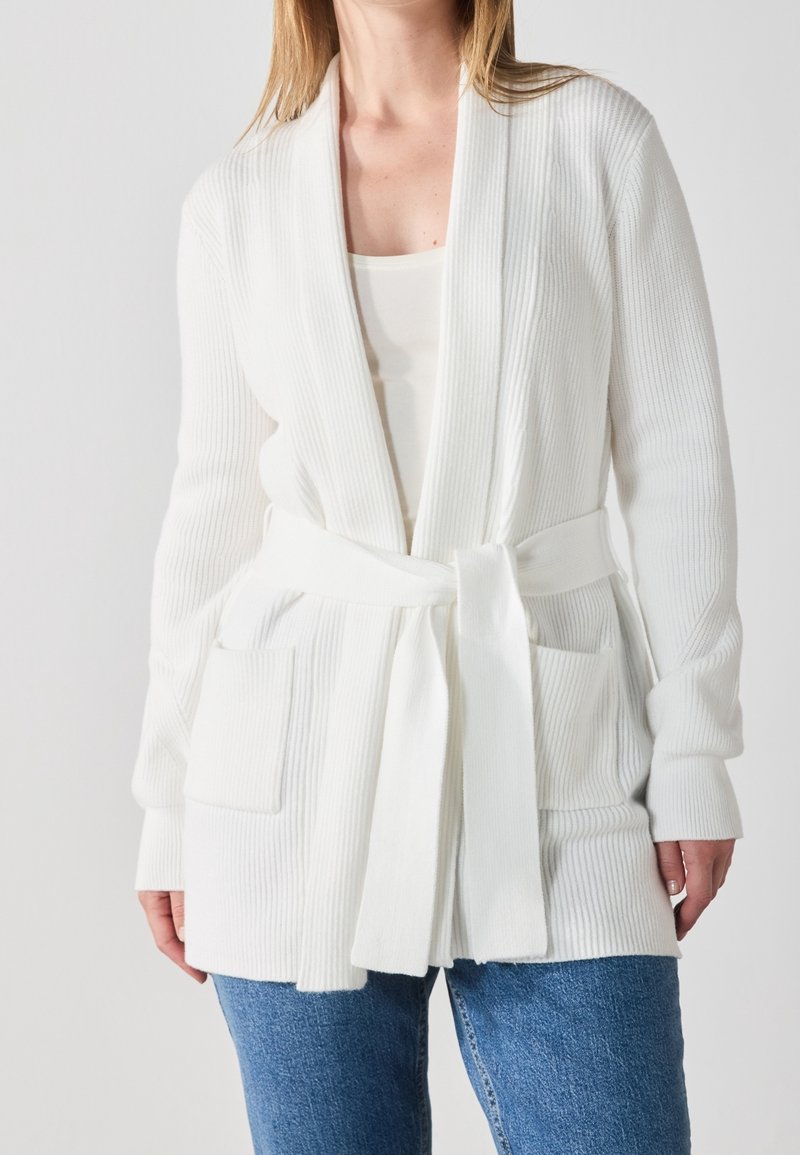 Femme portant un cardigan en maille côtelée blanche avec poches avant et ceinture nouée à la taille, associée à un jean bleu.