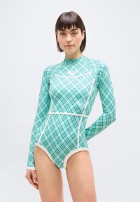 Γυναίκα που φοράει μαγιό με μακριά μανίκια, καρό σε πράσινο και μπλε χρώμα με λευκή επένδυση, στέκεται μπροστά σε ένα απλό φωτεινό φόντο.