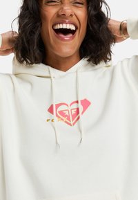 Sudadera con capucha color crema con un gran logo de corazón rosa y dorado. Presenta cordones y una tela ligeramente texturada.