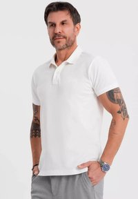 Vit bomullspiké med knäppning med tre knappar och korta ärmar. Har strukturerad tyg och en figursydd design, kombinerad med grå byxor.