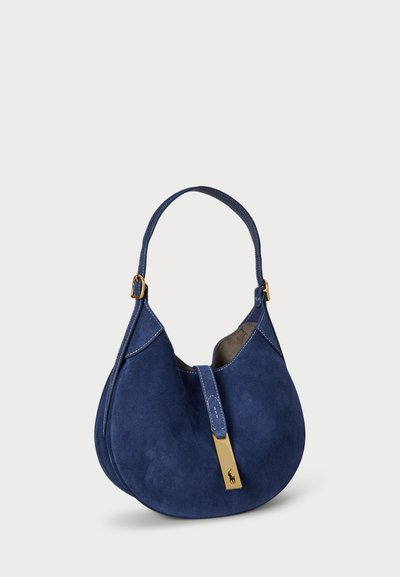 Navy blue suede shoulder bag with a curved shape, gold hardware accent, and stitched detailing on the sides and handle.