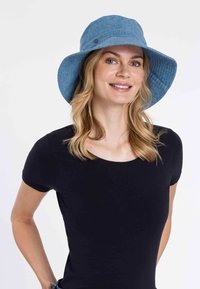 Denim kék vödörkalap széles karimával, texturált anyagból, és egy kis fémből készült logóval az oldalán. Egy nő fekete pólót visel.