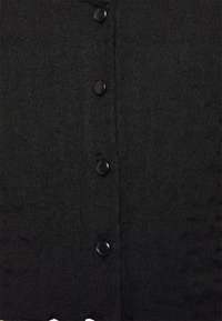 Svart texturerad tyg med ett vågigt mönster, med fem svarta knappar för stängning längs framsidan. Kanten är något vågig.