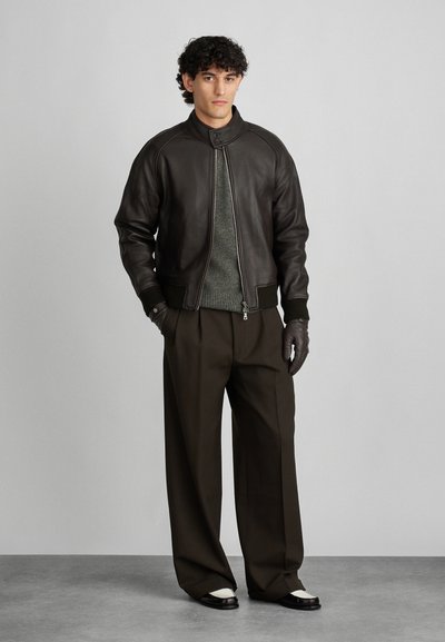 Καφέ δερμάτινο μπουφάν bomber με όρθιο γιακά, συνδυασμένο με πράσινο πουλόβερ, χύμα καφέ παντελόνια και σκούρα παπούτσια με λευκές λεπτομέρειες.