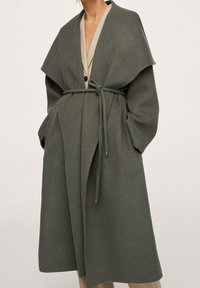 Lange, olijfgroene jas van zacht materiaal, met een brede kraag, ceintuur in de taille en zijzakken. De binnenzijde heeft een lichtbeige detail.