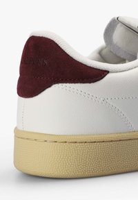 Zapatilla blanca de piel con un refuerzo en ante burdeos en el talón, suela de goma texturizada color crema y detalles de costura sutiles.