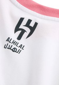 Camisa deportiva blanca con un ribete rosa en el cuello. Presenta un logo negro y el texto "AL HILAL" en árabe e inglés en la parte posterior. Material de algodón.