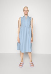 Vestido sem mangas azul claro com fecho de botão à frente, cintura franzida e comprimento até ao joelho. Combinado com sandálias chunky em lavanda.