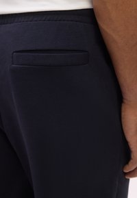 Κλείσιμο εικόνας από άτομο που φοράει σκούρο μπλε παντελόνι με πίσω τσέπη με φάσα και λευκό πουκάμισο που είναι φασκιωμένο μέσα στο παντελόνι.