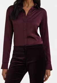 Burgunderfarbenes Satinhemd mit langen Ärmeln, spitzem Kragen, Knopfdetails und glatter Textur, kombiniert mit dunklen Samthosen.