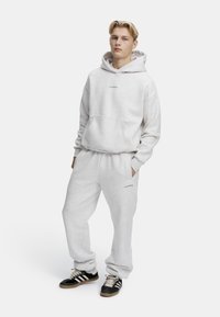 Grå sweatshirt og sweatsæt lavet af blødt stof, med en frontlomme og logo på brystet og låret. Sorte og hvide sneakers fuldender looket.